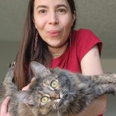 Image: N.K. Valek has light skin and long, straight brown hair. She wears a red shirt with long earrings of beaded red and white. Two choker necklaces decorate her neck. She purses her lips in an humored "oo" shape as her tortoiseshell maine coon cat twists in her arms to look at the camera with curiosity.